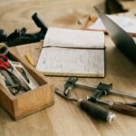 A creative woodworking workspace with tools, notebook, and laptop on a wooden table.